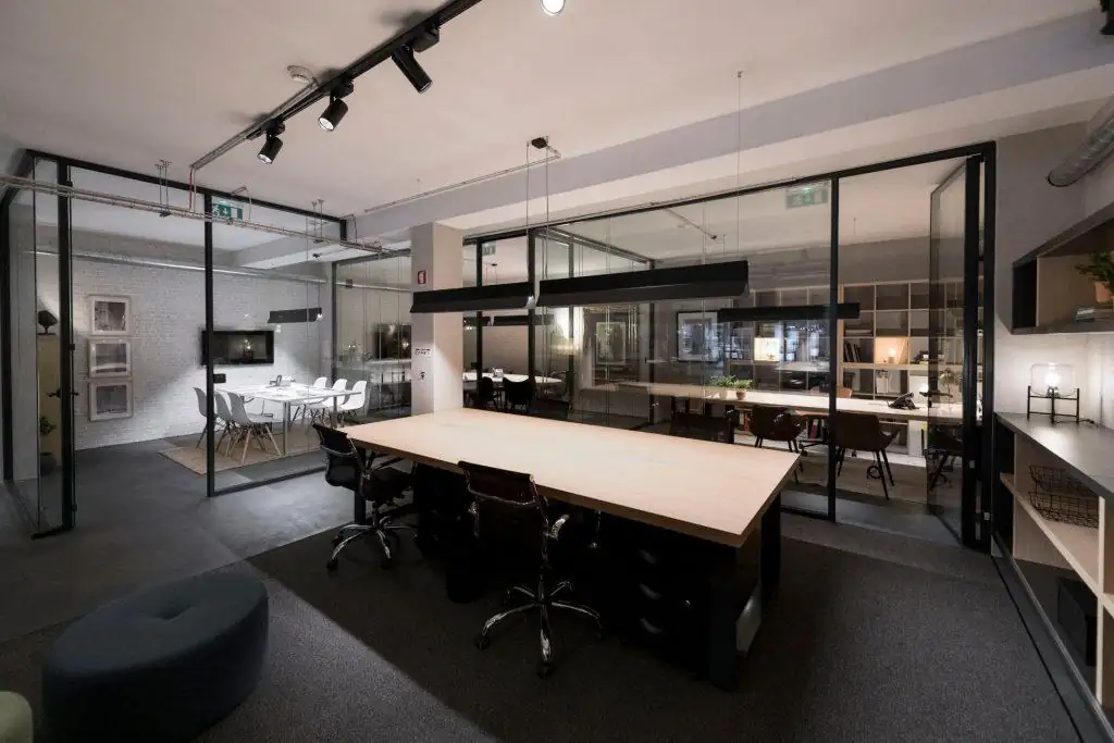 Vista de uma mesa de trabalho em open space no Outglocal Coworking Aveiro, com gabinetes privados com divisórias em vidro ao fundo.