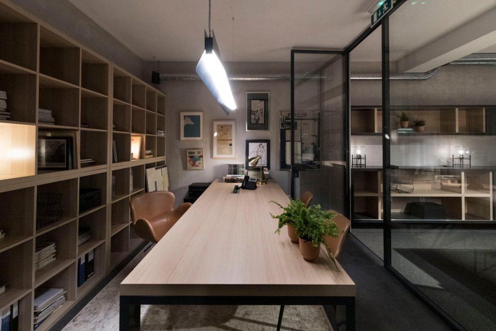 Zona de trabalho comum no Outglocal Coworking Aveiro com uma longa mesa de madeira, uma planta e uma estante de livros.