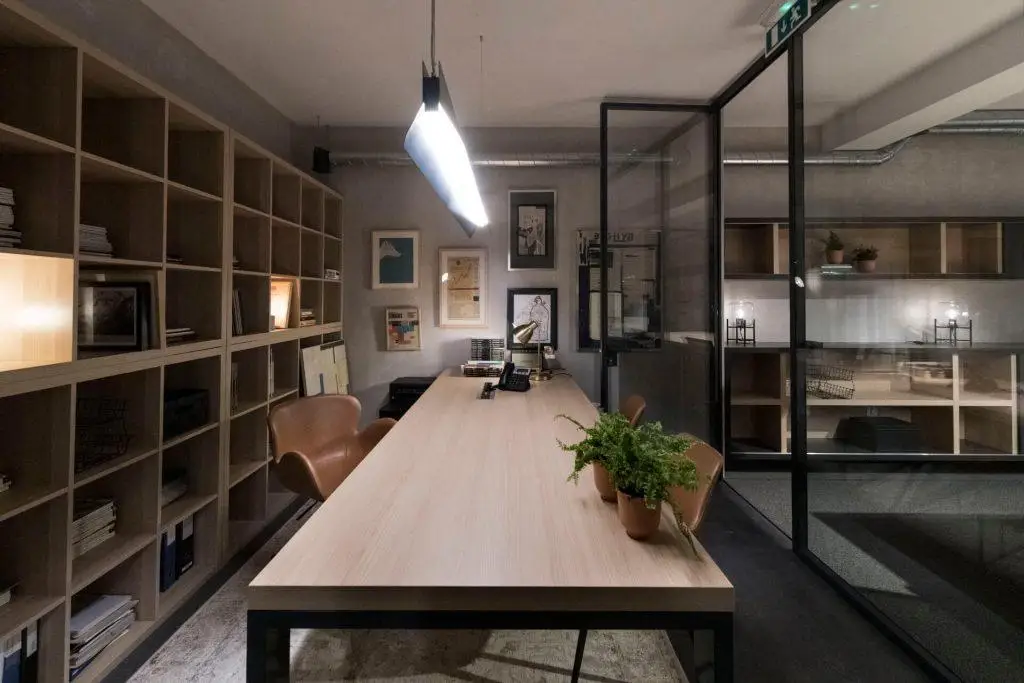 Zona de trabalho comum no Outglocal Coworking Aveiro com uma longa mesa de madeira, uma planta e uma estante de livros.